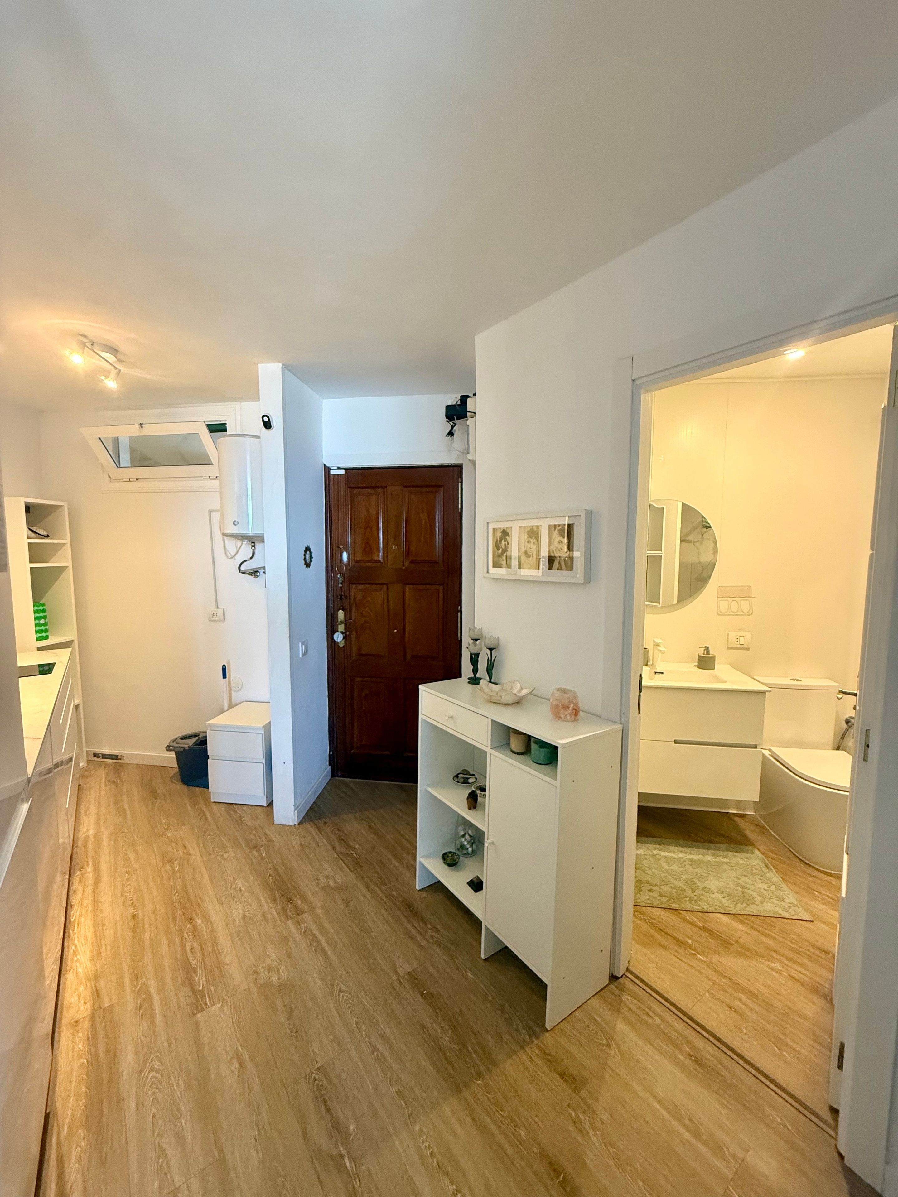 Un pasillo que conduce a un baño con lavamanos, espejo y retrete blancos. Hay una puerta de madera, un estante blanco y un pequeño cubo de basura.