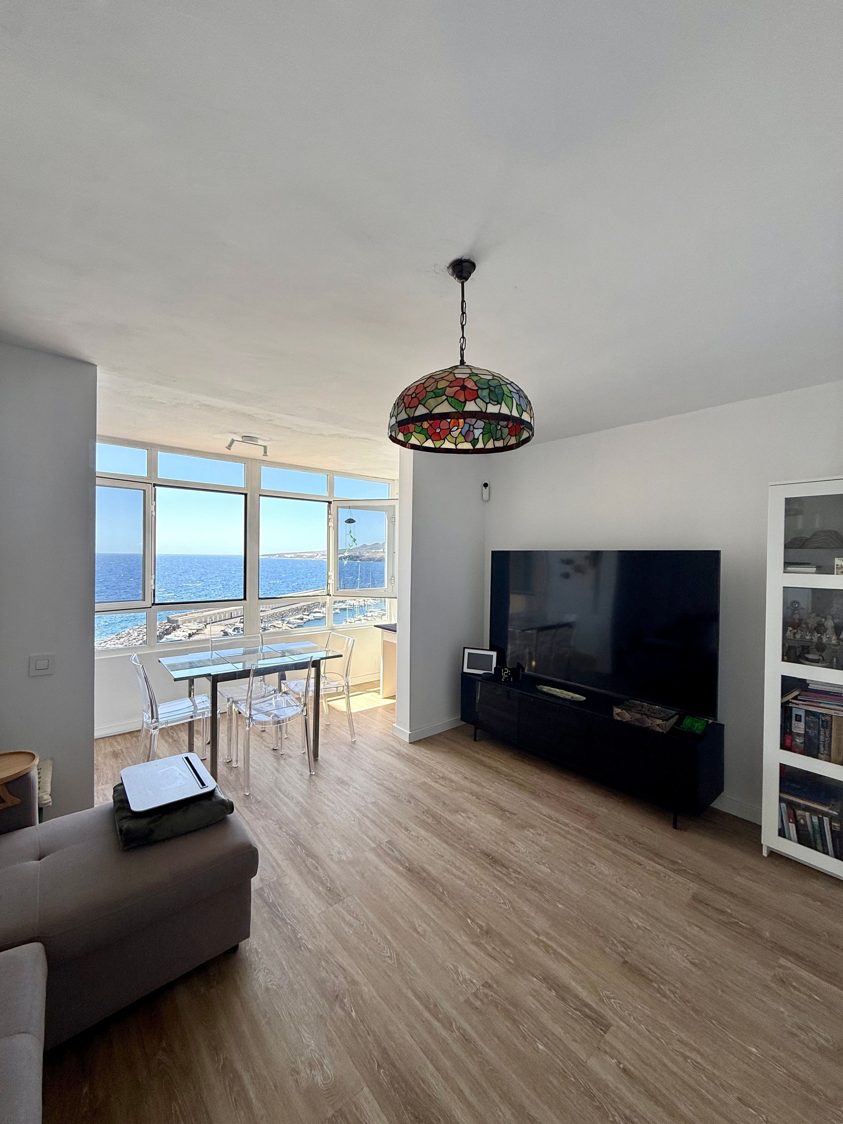 Een moderne woonkamer met een grote televisie, een eethoek met stoelen en uitzicht op de zee door grote ramen.