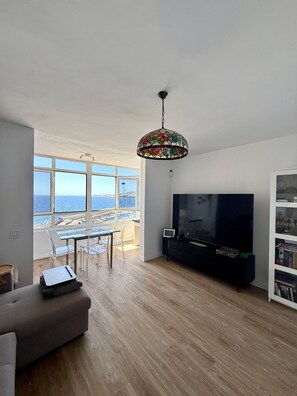 A modern living room with a large TV, a dining area with chairs, and a view of the sea through large windows.
