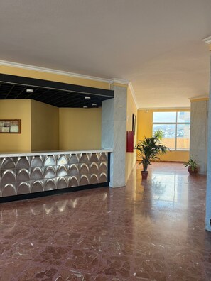 Um hall amplo com piso de mármore, balcão de recepção com painel decorativo e plantas em vasos.
