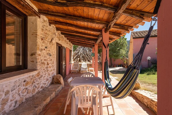 Un porche cubierto con una mesa y sillas, una hamaca y un muro de piedra.