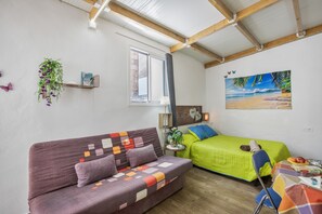 A room with a sofa, a bed, a table with fruits, a painting of a beach, and a window with curtains.