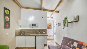 Eine kleine Küche mit Mikrowelle, Spüle und einem Kühlschrank. Ein Sofa mit einem farbenfrohen Kissen und ein grüner Stuhl sind ebenfalls vorhanden.
