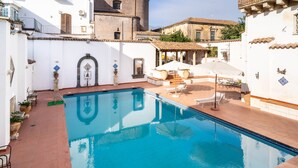 Piscina com deck ladrilhado, área de estar ao ar livre e um edifício branco com azulejos decorativos e uma porta.