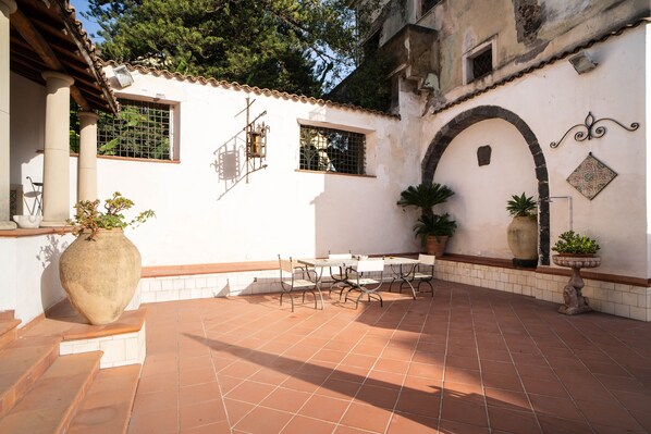 Une cour dallée de carreaux de terre cuite, un mur blanc avec une base carrelée, une grande plante en pot, une table avec des chaises et une statue en pierre.