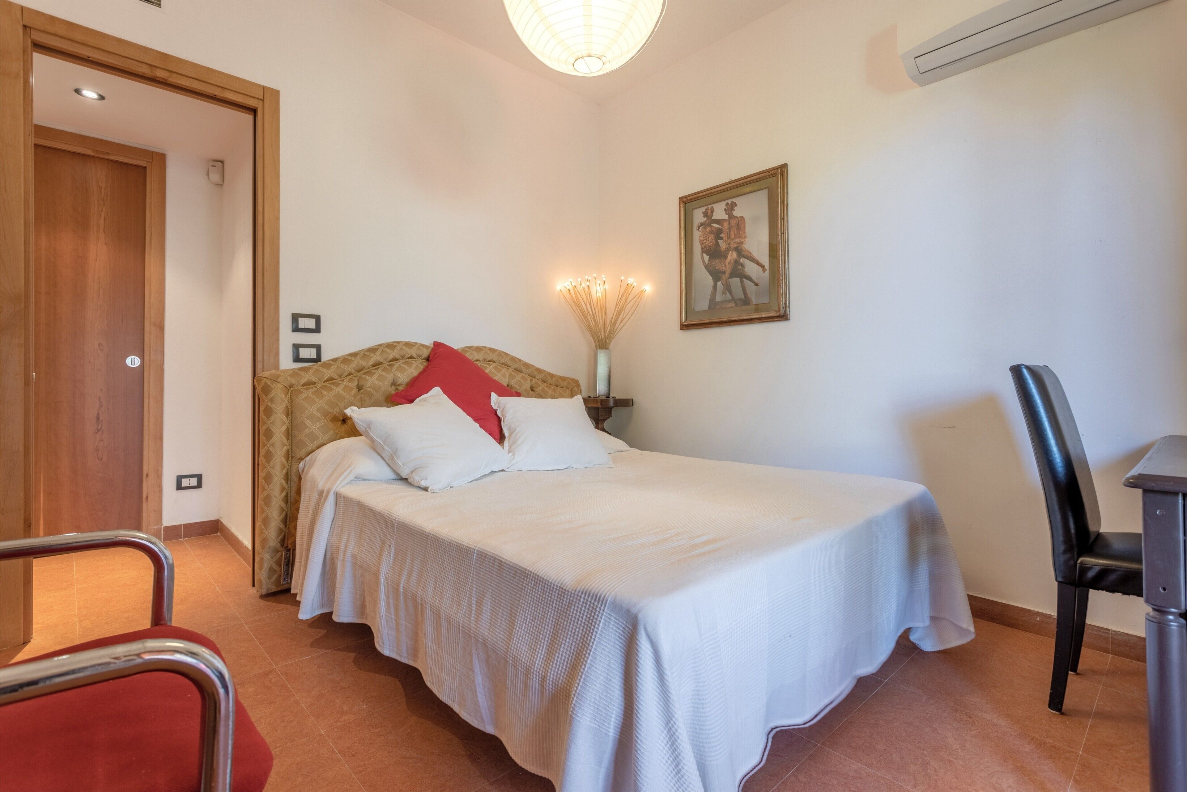 A bedroom with a bed, a chair, a table, a door, and a wall-mounted air conditioner.