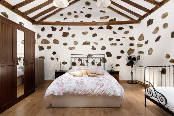 A bedroom with a stone wall, wooden ceiling beams, a bed with floral bedding, bedside tables, and a floor lamp.