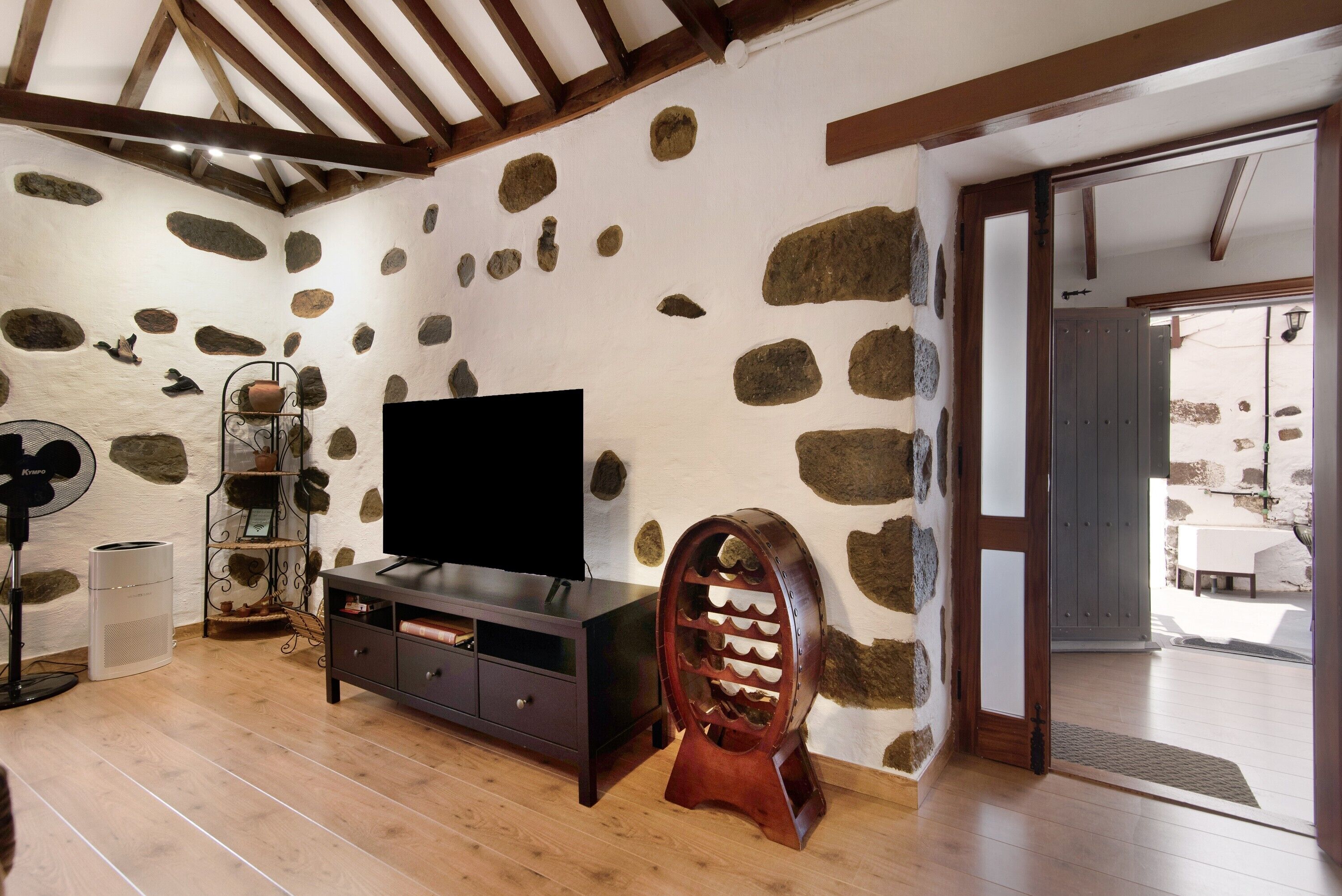 Une pièce avec un mur en pierre, un téléviseur posé sur un meuble en bois, un ventilateur et une porte donnant sur l’extérieur.