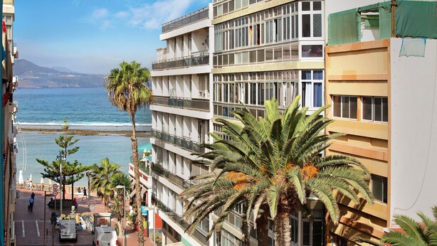 A coastal building with balconies overlooking the sea, palm trees, and a clear sky.
