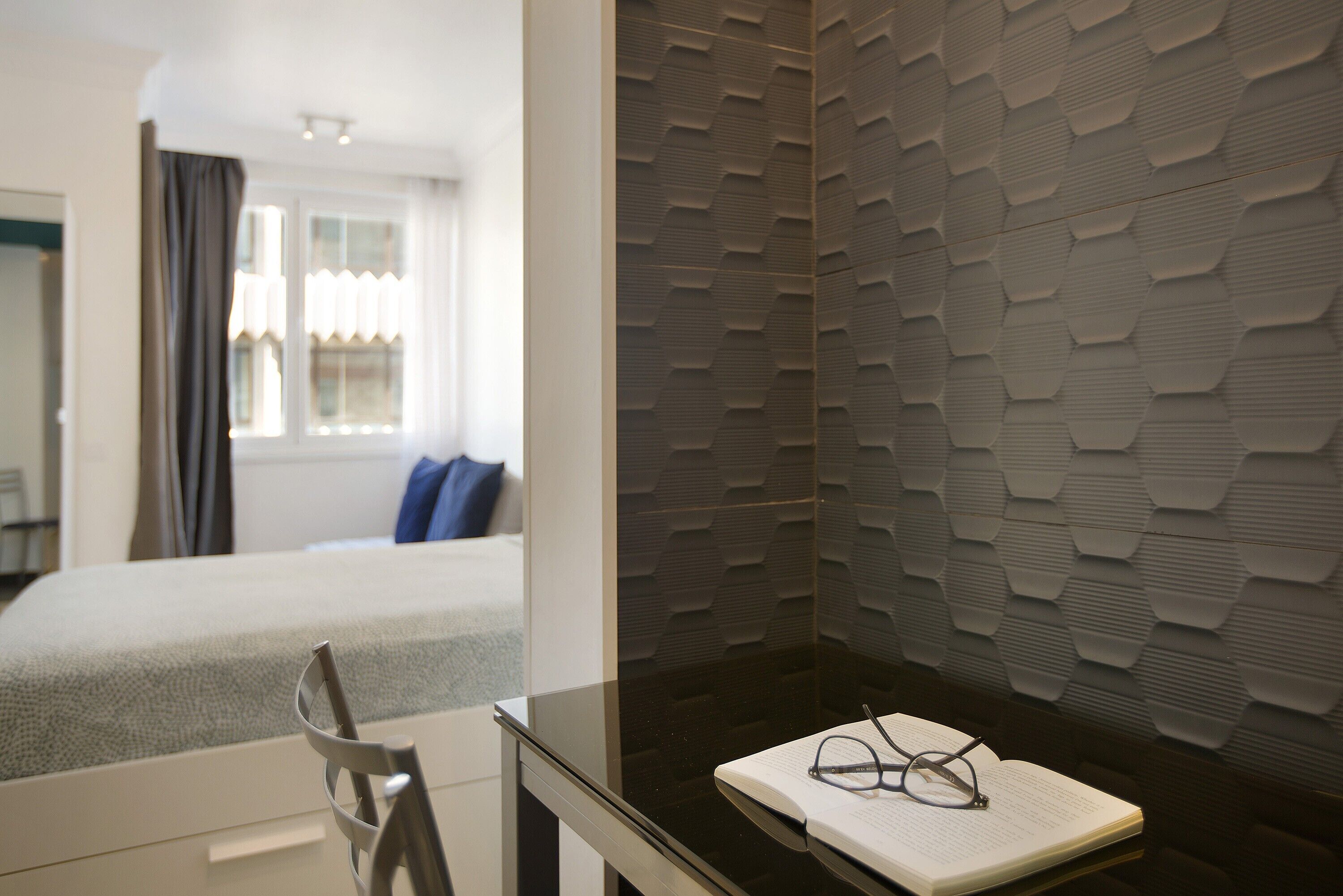 Habitación de hotel con cama, escritorio con silla, ventana con cortinas y pared con textura.