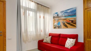 A room with a red sofa, a wooden bench, a painting of boats on a beach, and a window with sheer curtains.