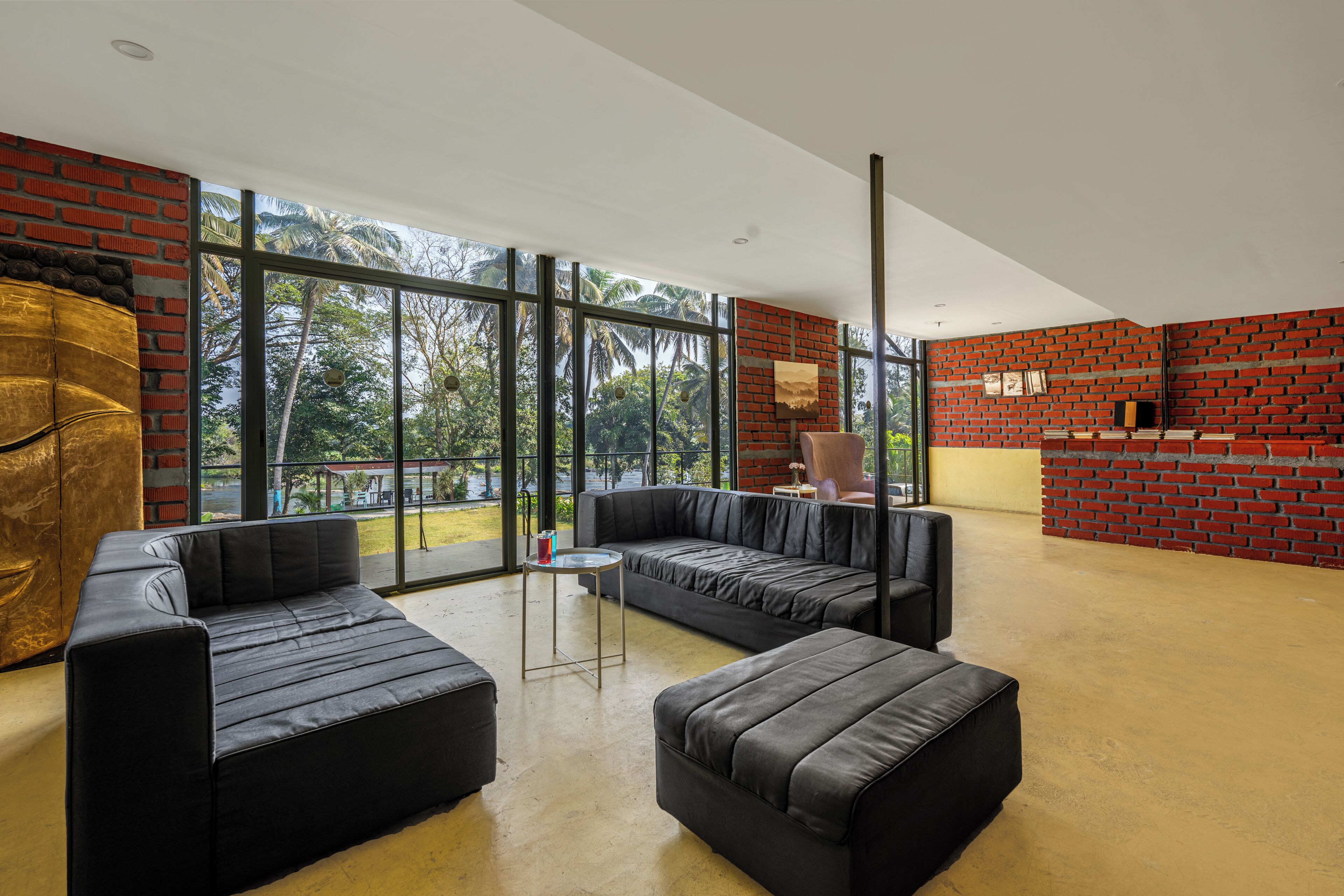 Ein modernes Wohnzimmer mit schwarzen Ledersofas, einem kleinen Couchtisch und einer Ziegelwand als Blickfang. Große Fenster bieten Blick auf Palmen und ein Gebäude im Freien.