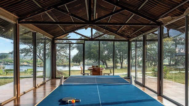 An indoor ping pong table with a view of a park and a body of water through large windows. - ATMAVEDA RIVERSIDE HOMESTAY (Shrirangapattana)