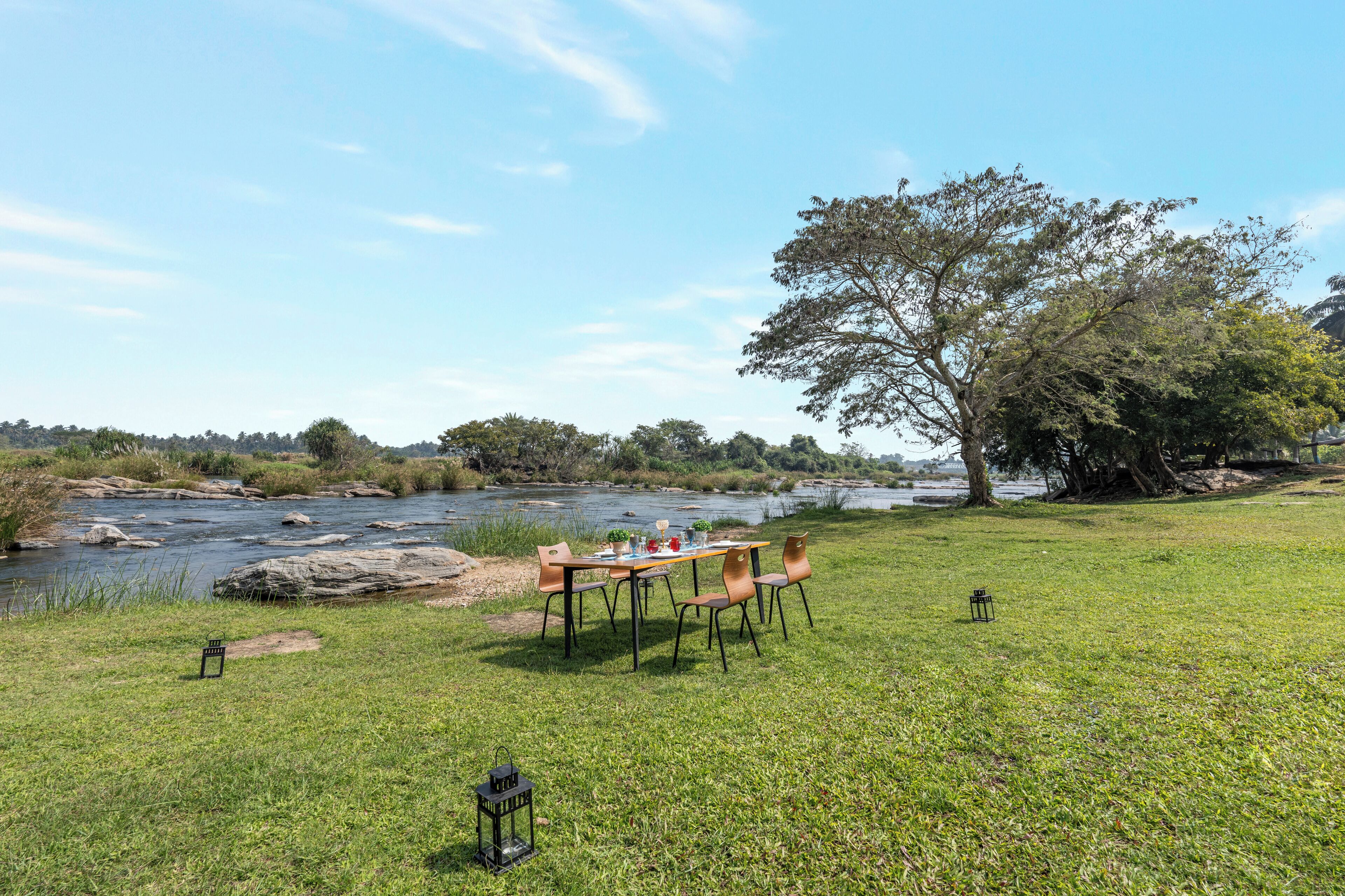 An outdoor dining area with a table and chairs by a river, surrounded by grass and trees.