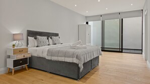 Une chambre à coucher moderne avec un lit gris, des tables de chevet blanches, une grande fenêtre et une armoire.
