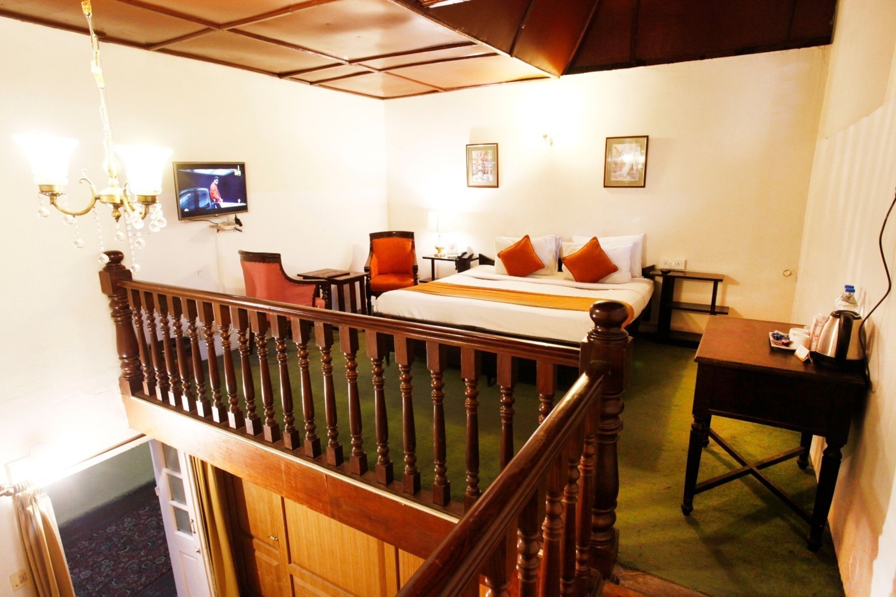 Quarto de hotel com escada de madeira, cama com almofadas laranja, escrivaninha com chaleira e televisão na parede.