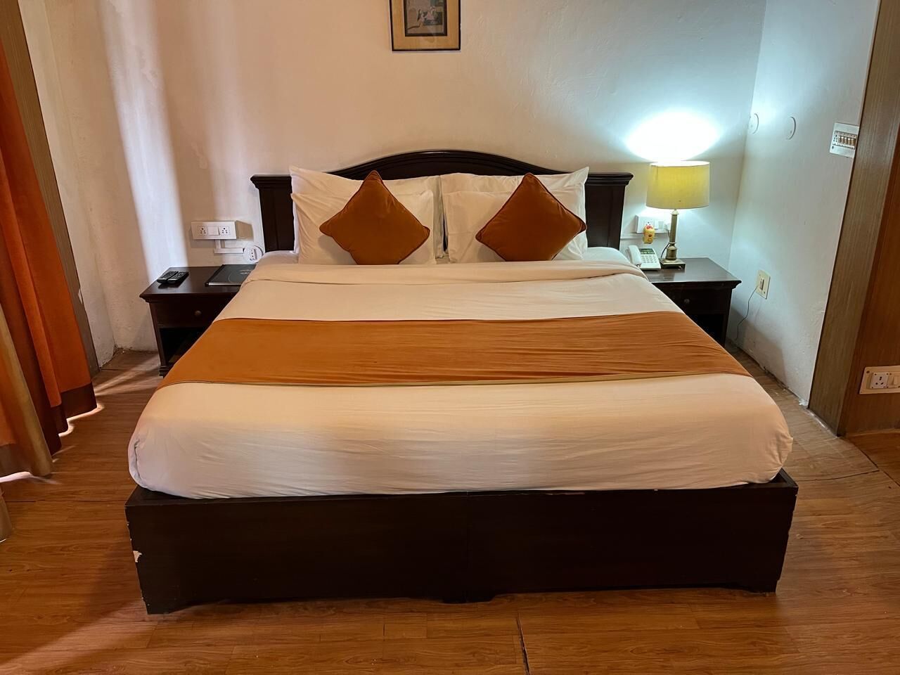 Un lit bien fait, avec une tête de lit sombre, une literie blanche et deux oreillers marron. On y trouve une table de chevet avec une lampe et un téléphone. La pièce est dotée d’un plancher en bois et une porte se trouve sur le côté gauche.