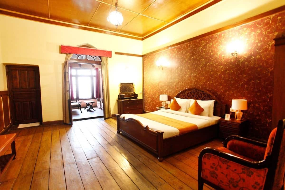 Habitación de hotel con piso de madera, una cama grande con ropa de cama blanca y naranja, un cabecero de madera, dos mesitas de noche con lámparas, un sillón tapizado rojo y una puerta de madera.