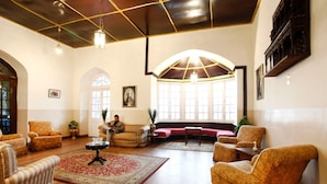 Un salon spacieux doté d’un haut plafond, d’un plancher en bois et d’une grande fenêtre qui laisse entrer la lumière naturelle. On y trouve plusieurs fauteuils et un canapé disposés autour d’une table basse centrale.