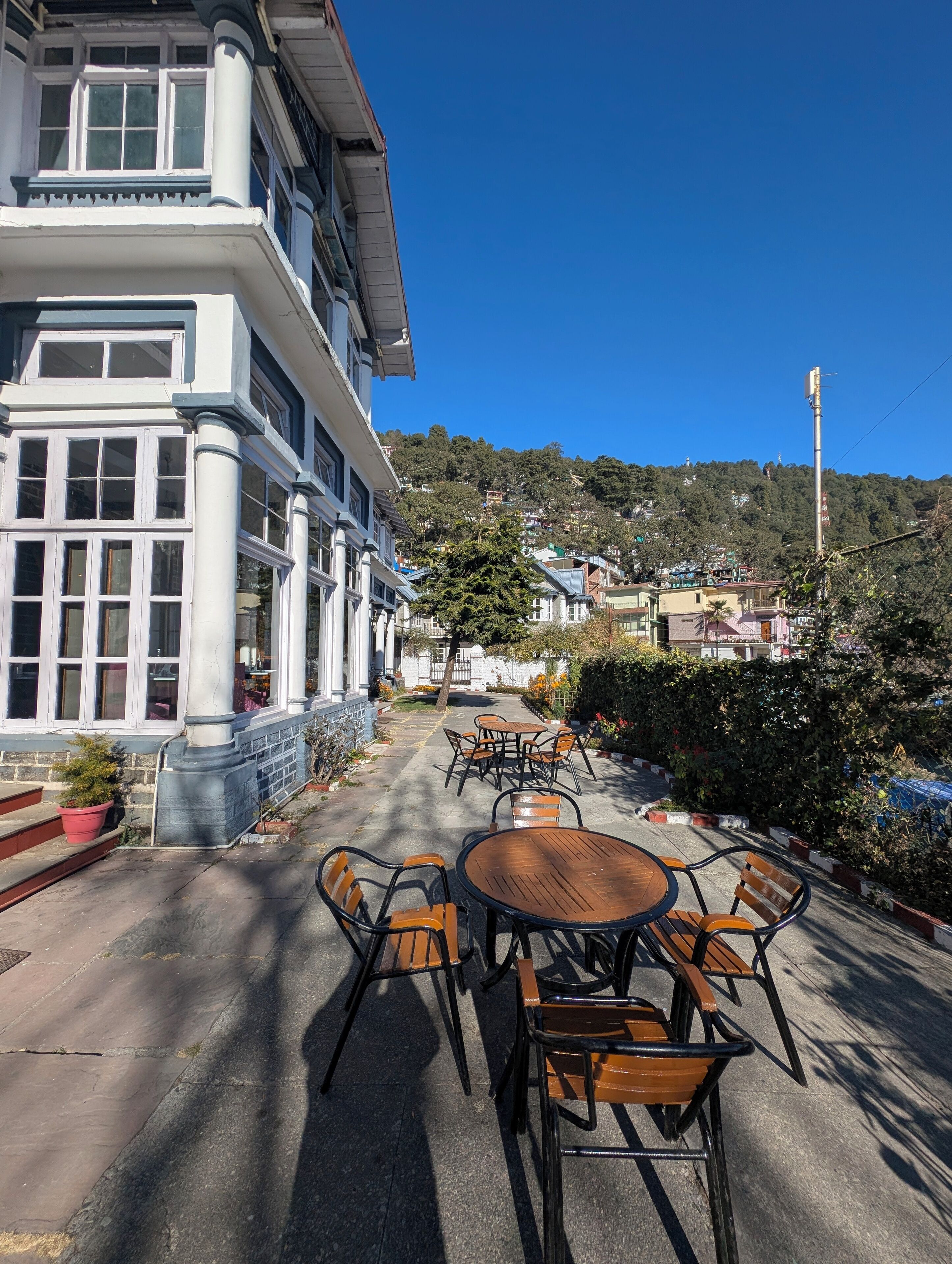 Un edificio con grandes ventanales y un balcón, zona de estar al aire libre con mesas y sillas, y vistas a una colina con casas.
