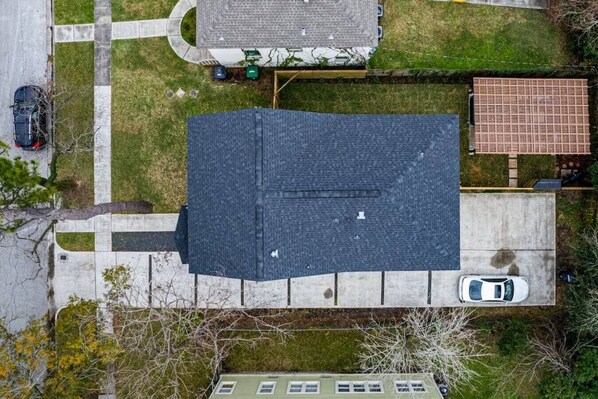 An aerial view of a residential area with a house featuring a dark roof, a driveway, and a neighboring house with a red roof.