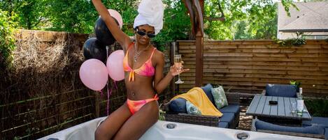 A person in a bikini sitting in a hot tub with balloons and a drink, surrounded by outdoor furniture and a wooden pergola.
