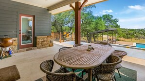 Un patio couvert avec une table et des chaises, un ventilateur de plafond et une vue sur une piscine et des arbres.
