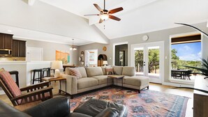 Un vaste salon comprenant un grand canapé d’angle, un fauteuil en cuir et un tapis à motifs. On y trouve également un ventilateur de plafond, un coin repas avec une table et des chaises, ainsi qu’une cuisine équipée de placards et d’un four à micro-ondes.