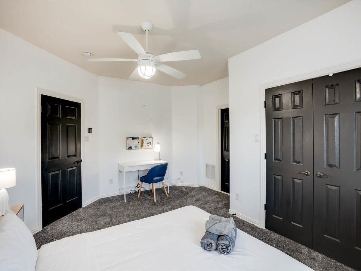 Une chambre à coucher comprenant un lit, un bureau, une chaise, un ventilateur de plafond et deux portes.