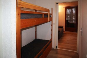 A bunk bed with a wooden frame and a mattress, a door leading to a room with a bed, and a window with a view of trees.