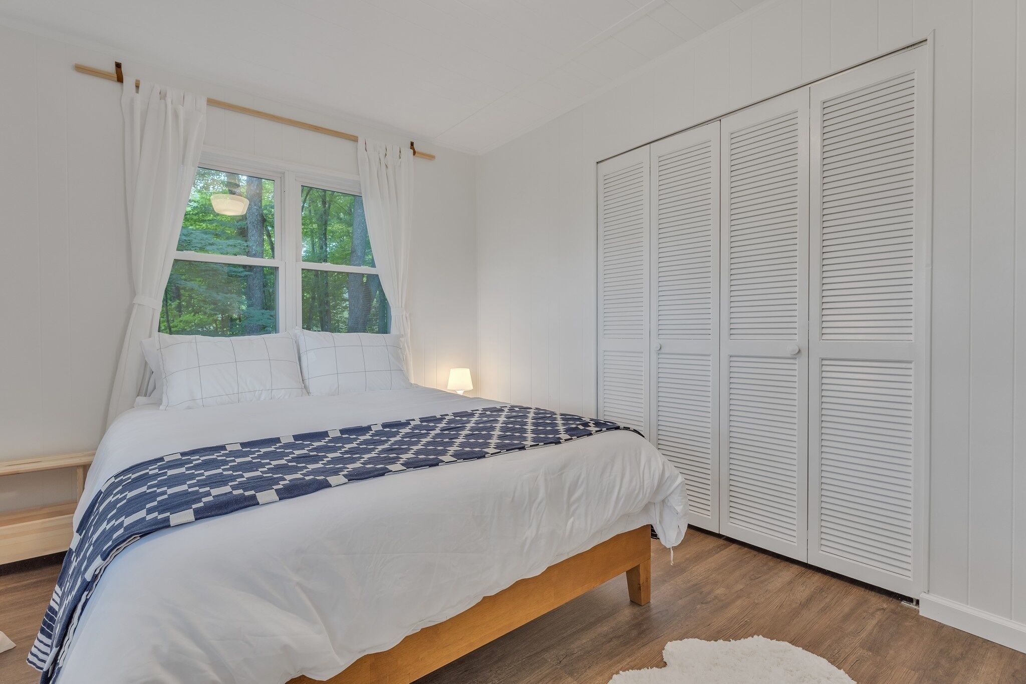 Une chambre à coucher avec un lit en bois, des rideaux blancs, une fenêtre donnant sur des arbres et une armoire blanche.