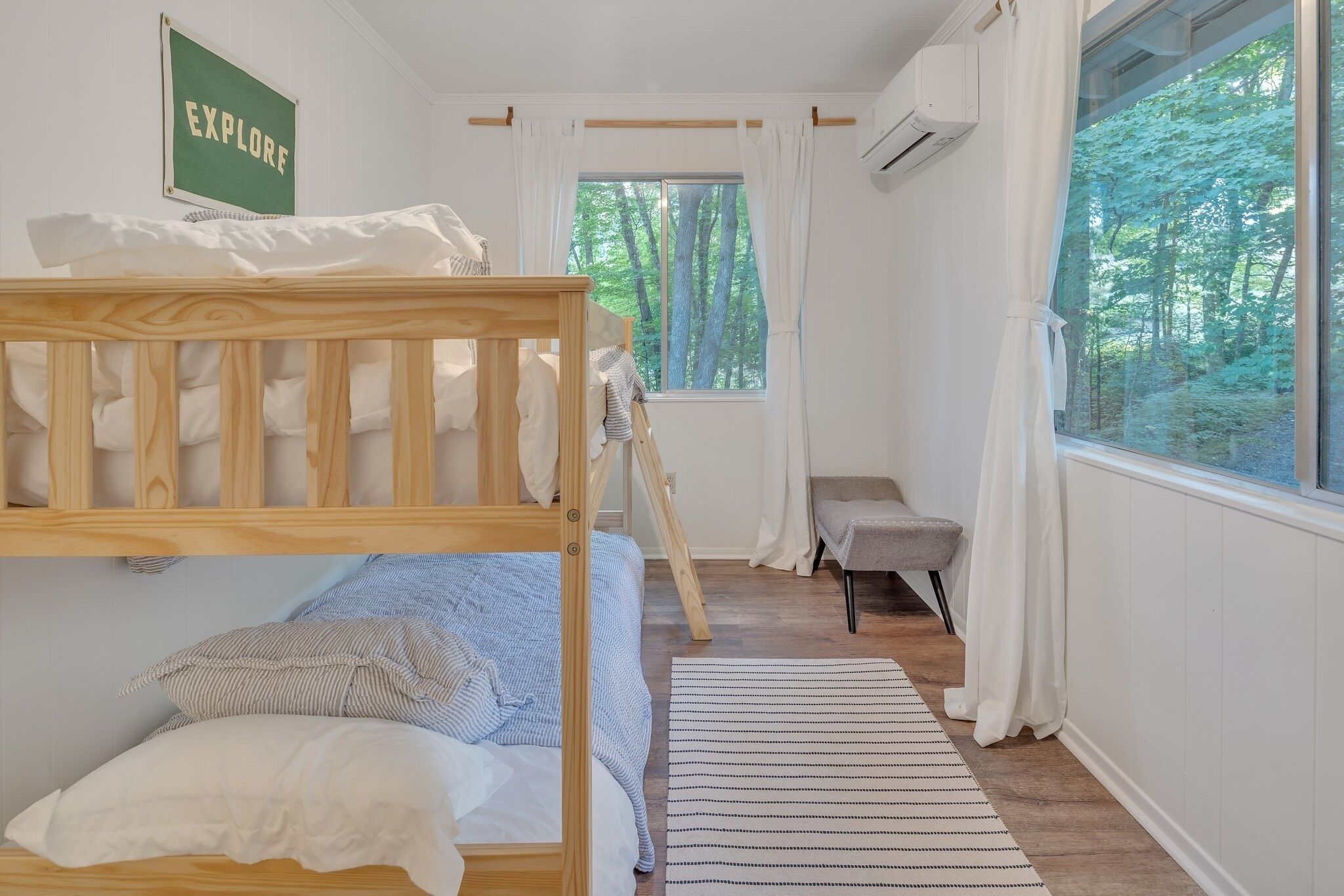 A bedroom with a bunk bed, a single bed, a window with a forest view, a small chair, and a sign that says "EXPLORE".