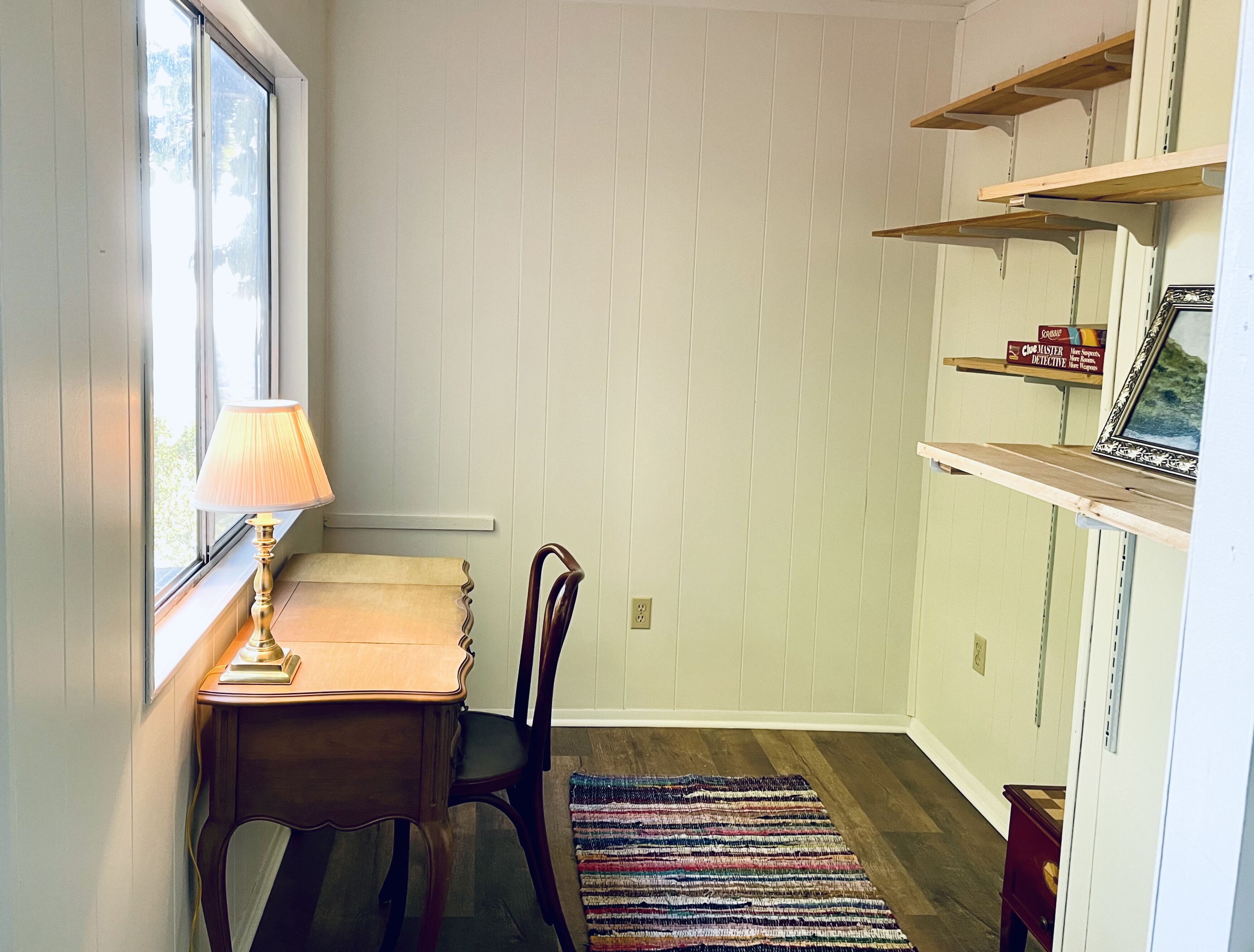 Ein Zimmer mit einem Holztisch, einem Stuhl, einer Lampe, einem farbenfrohen Teppich und Regalen mit Büchern und einem gerahmten Bild.