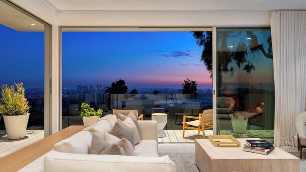 A modern living room with a white sofa, a wooden coffee table, and a glass door leading to a balcony with a city view at dusk.
