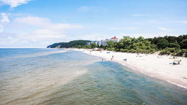 A beach with clear waters, a sandy shore, and a building with a red roof in the background. - Rezydencja Korab (Międzyzdroje)