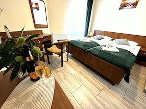 Una habitación de hotel con una cama, una silla, un escritorio, un radiador, una planta y una botella de champán.