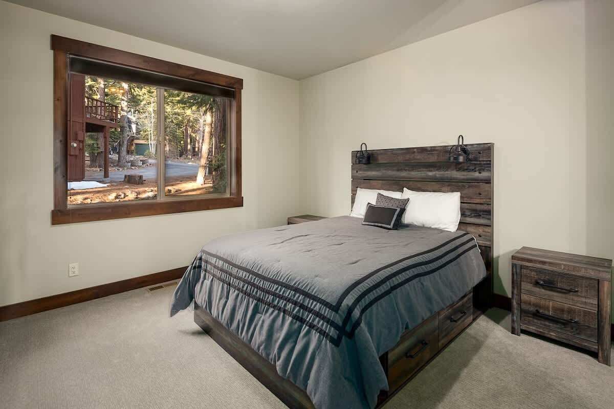 Een slaapkamer met een groot raam, een bed met houten hoofdeinde, een nachtkastje en een klein houten dressoir.