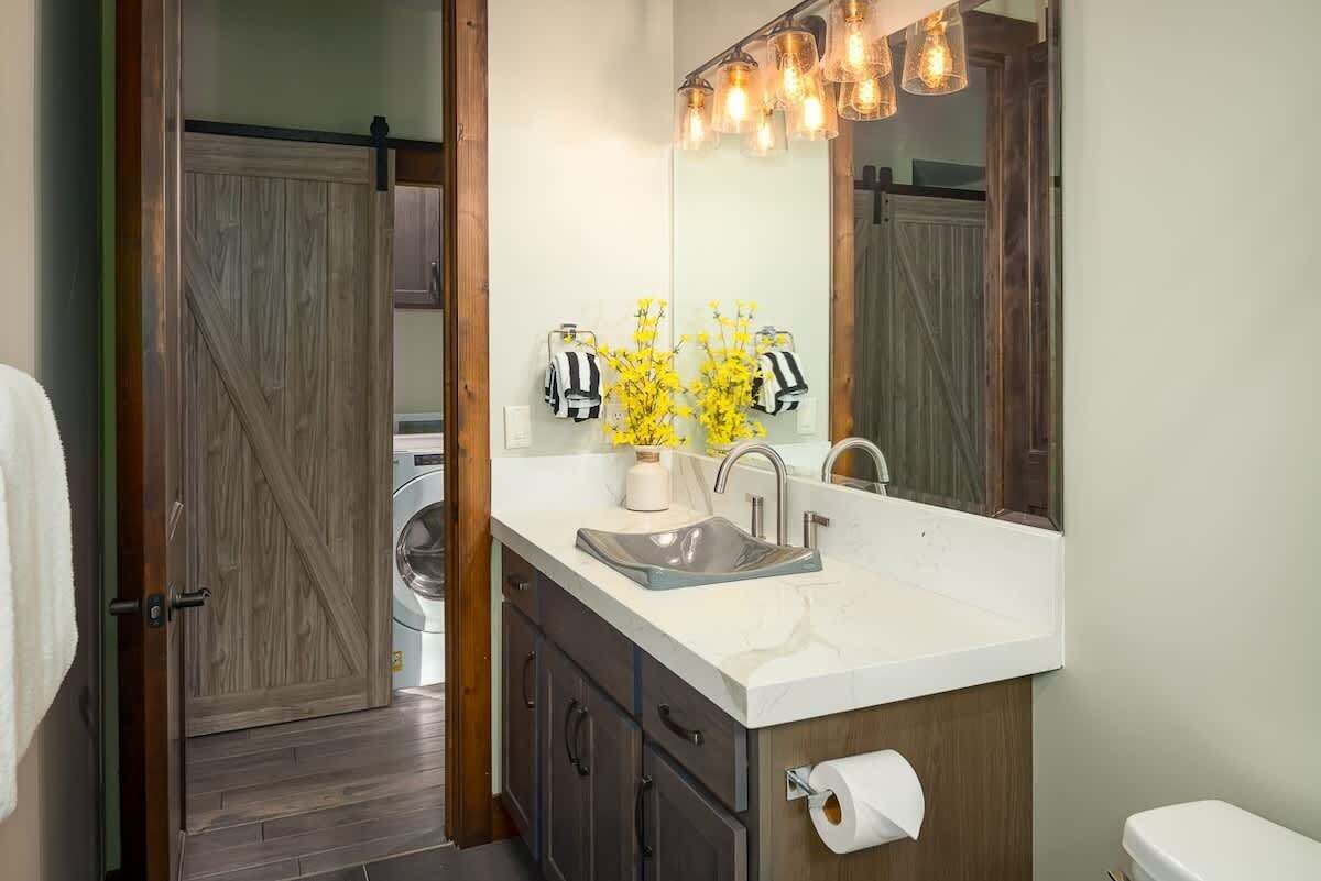 Un baño con puerta de madera, un lavabo con espejo, un inodoro y una lavadora al fondo.