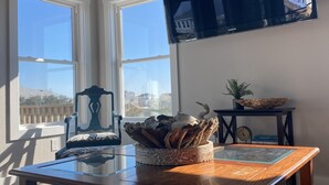 Una habitación con un escritorio de madera, un televisor montado en la pared, una ventana con vistas a edificios y una mesita con una planta y un reloj.