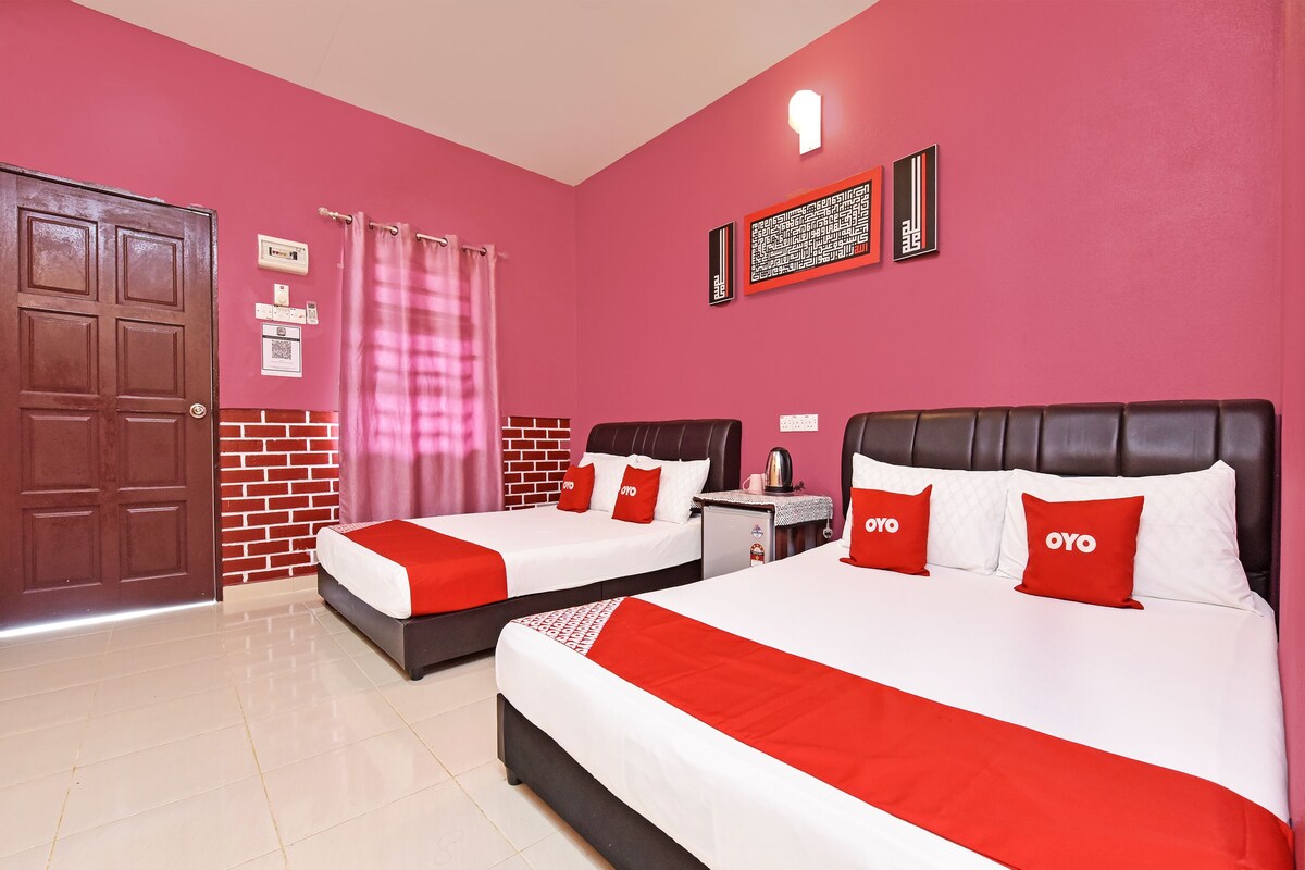 A hotel room with two beds, each with a red and white bedspread and matching pillows. There is a small table with a kettle and a cup, and a wall-mounted light fixture above the beds.