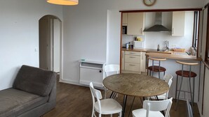 Un espace de vie compact comprenant un canapé, une table ronde en bois avec des chaises, un coin cuisine avec des placards et une horloge, ainsi qu’un luminaire suspendu au plafond.