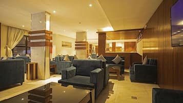 A modern hotel lobby with a central seating area, a bar, and a television screen on the wall.