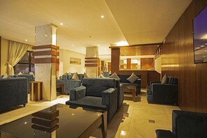 A modern hotel lobby with a central seating area, a bar, and a television screen on the wall.