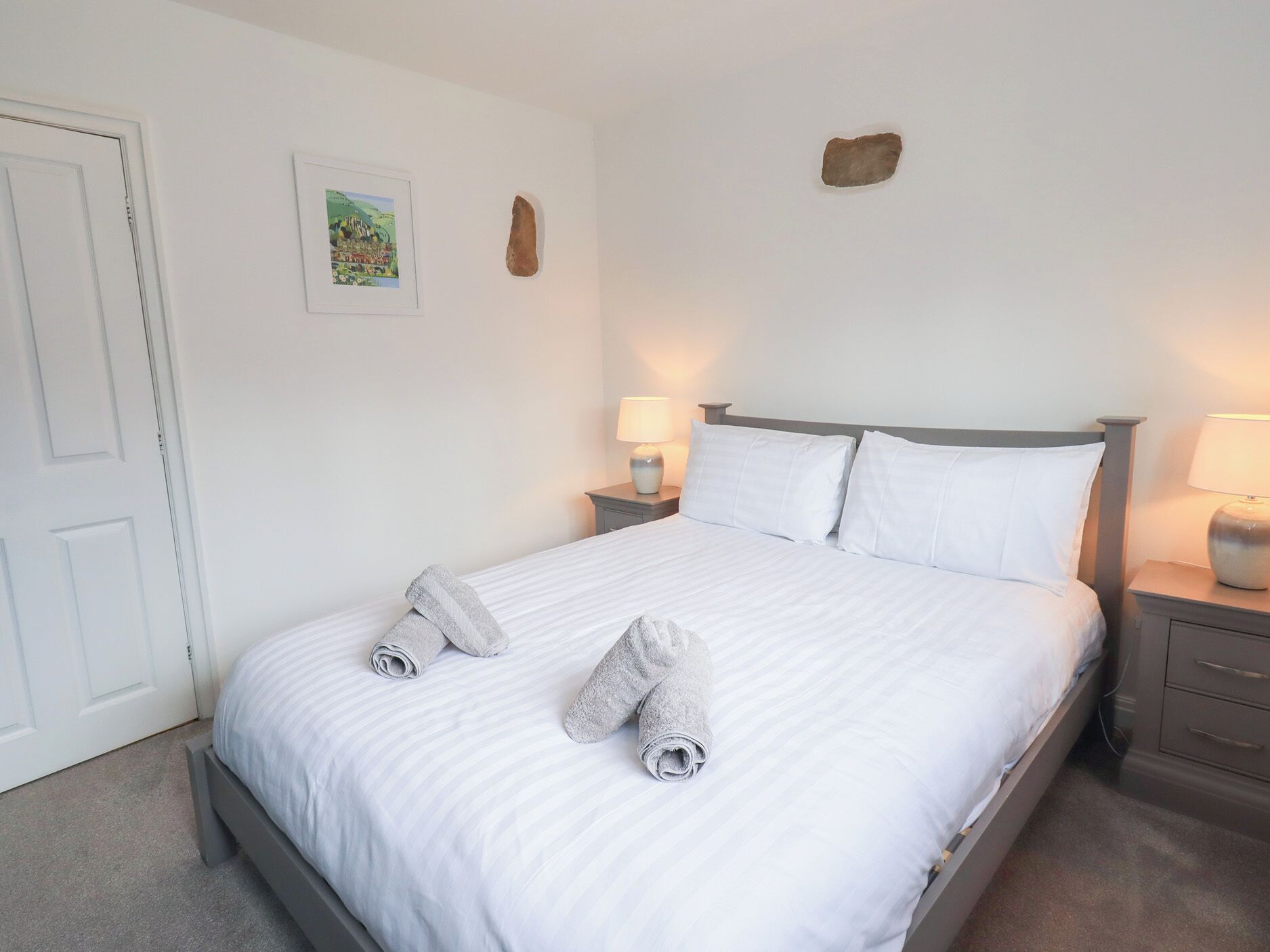 Un letto rifatto con lenzuola bianche e due asciugamani arrotolati, un quadro appeso al muro e una piccola decorazione in pietra.