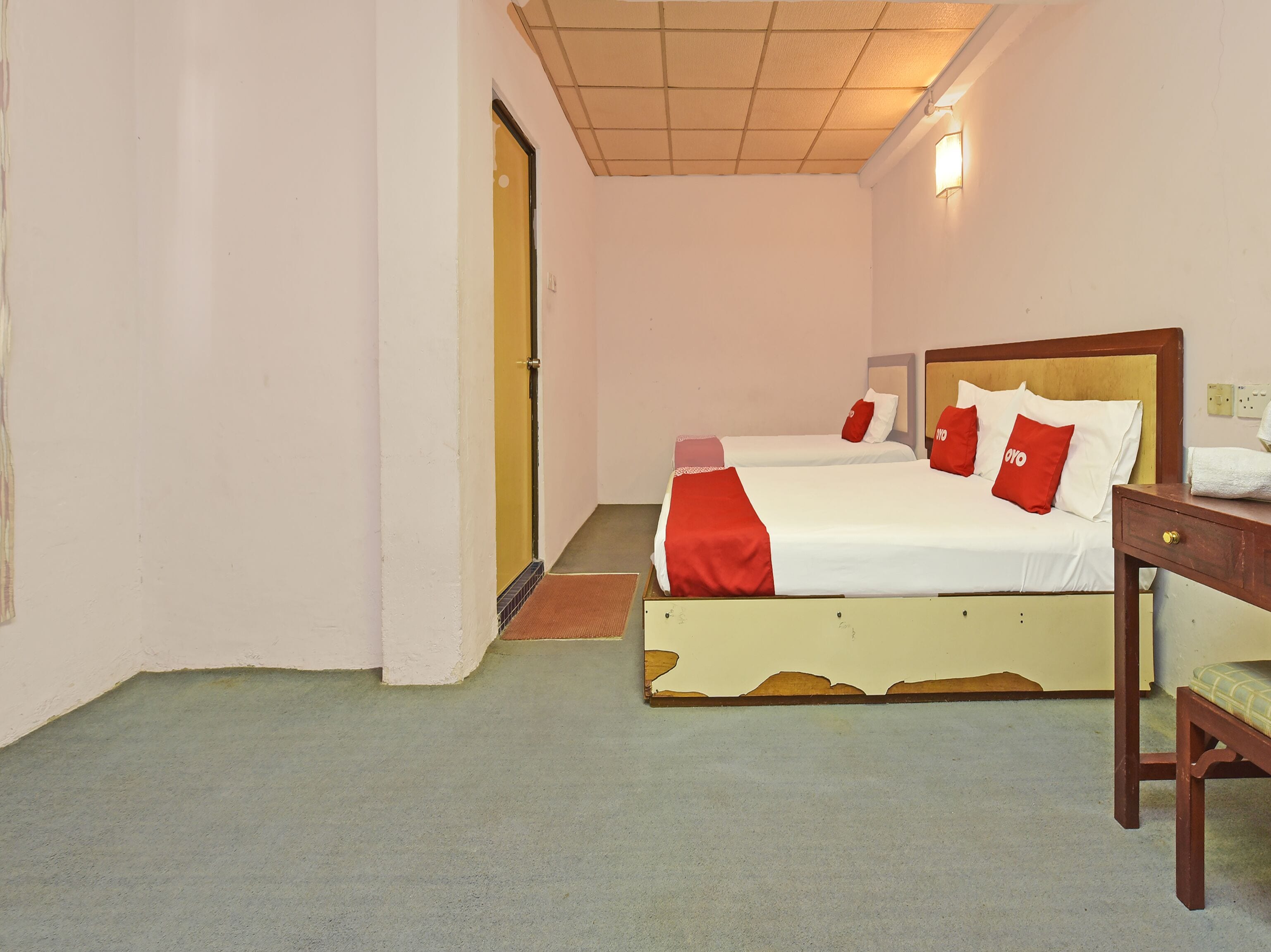 A hotel room with two beds, a wooden nightstand, a bench, and a door leading to another room.