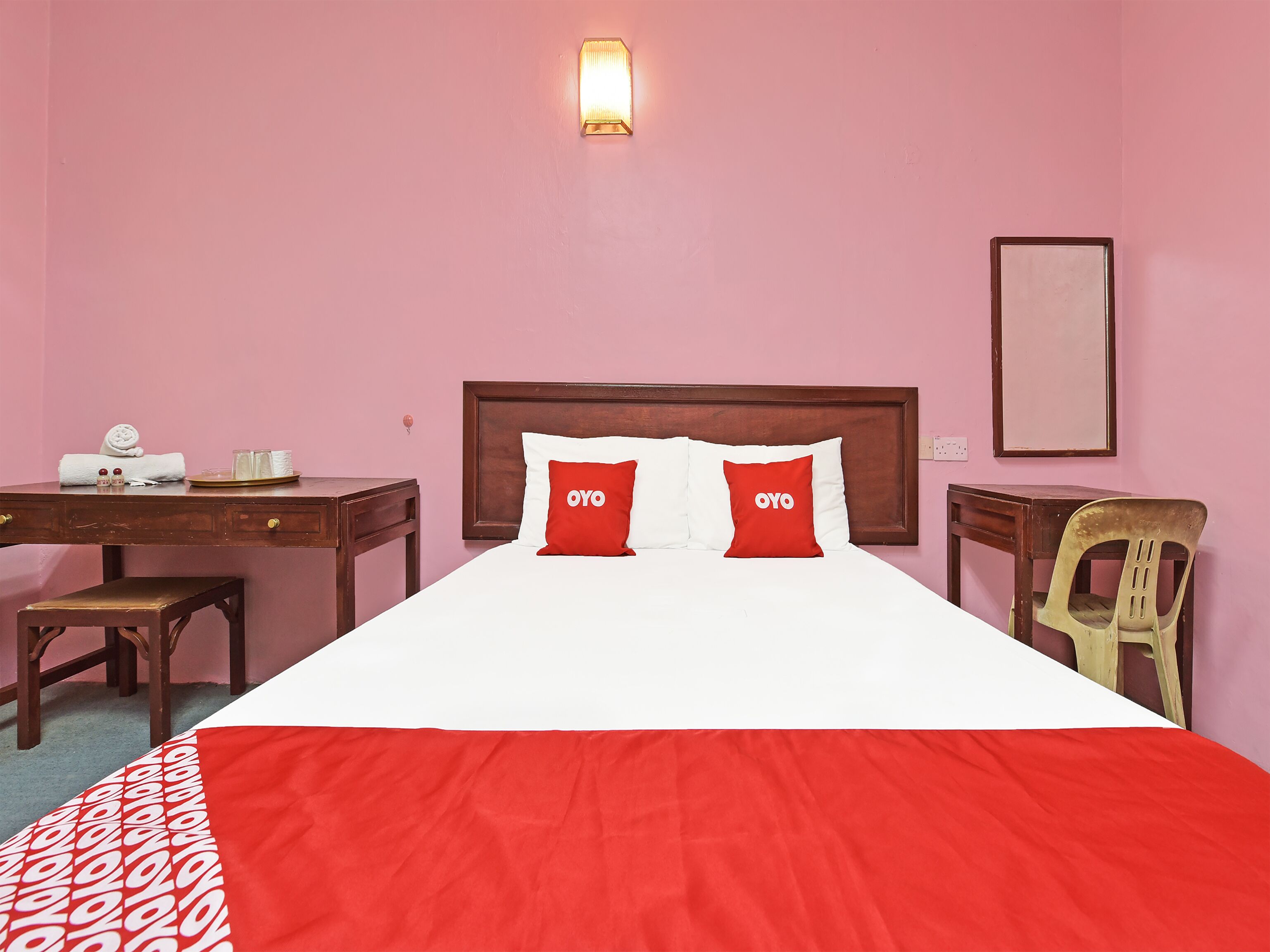 Une chambre d’hôtel avec un lit en bois, deux oreillers rouges portant la marque « OYO », une table de chevet en bois, un bureau avec une chaise et un luminaire fixé au mur.