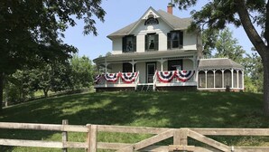 A two-story house with a porch decorated with red, white, and blue banners, a covered porch area, and a wooden fence in the foreground. - The Lodge at the Ranch - secluded but close to Galena and golf. (Elizabeth)