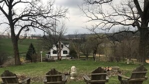A rustic outdoor seating area with wooden Adirondack chairs arranged around a fire pit, set against a backdrop of a white house and open fields. - The Lodge at the Ranch - secluded but close to Galena and golf. (Elizabeth)
