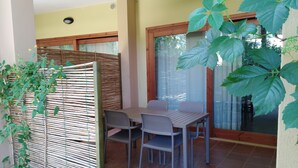 A patio with a table and chairs, a bamboo fence, and a large window with curtains. - two-room apartment with pool 200mt from the sea (Santa Margherita di Pula)
