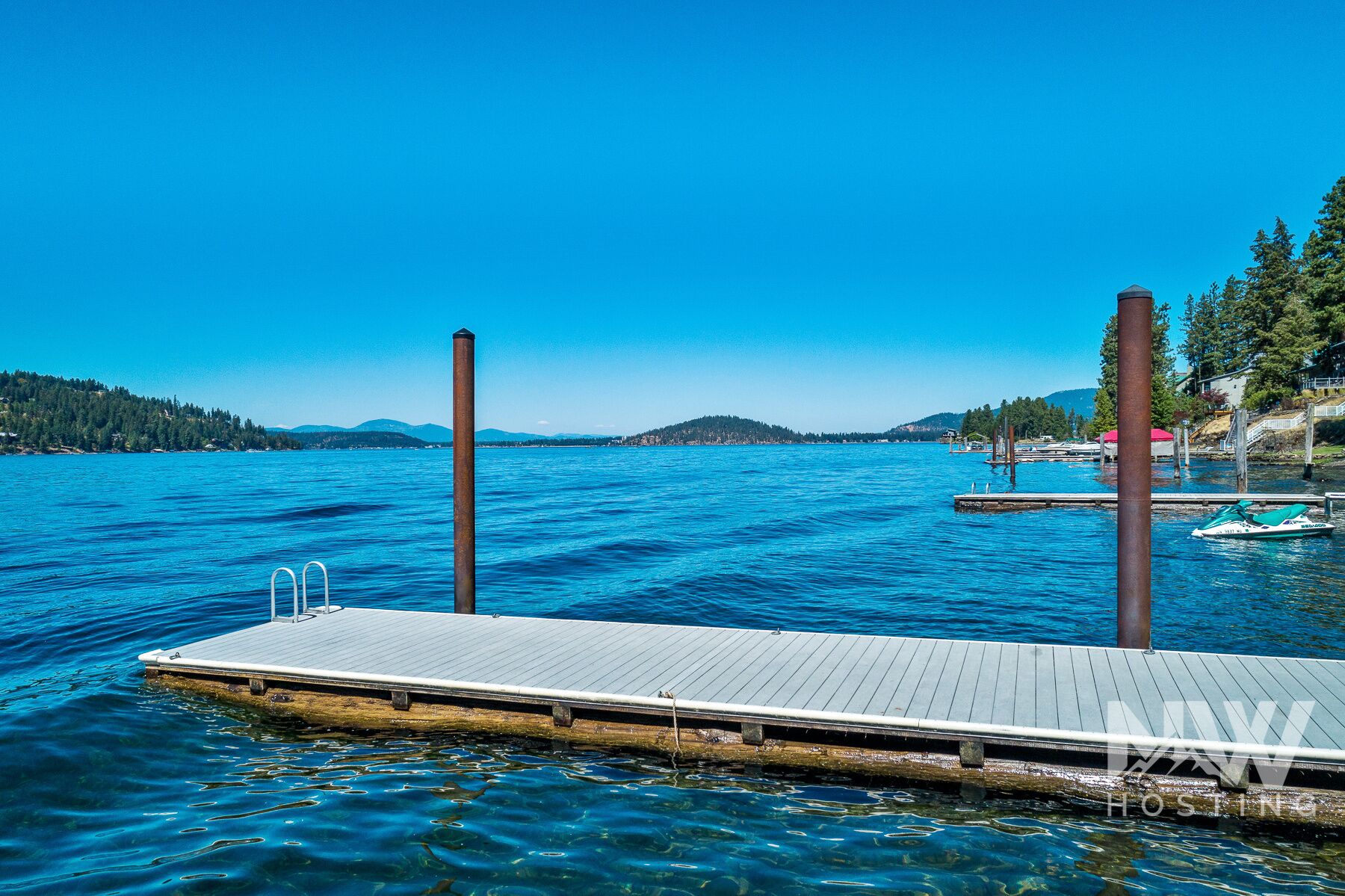 A wooden dock extending into a calm lake with clear blue water, surrounded by hills and trees.